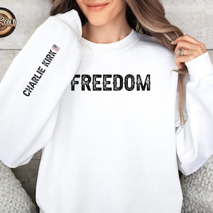 May include: White sweatshirt with the word "FREEDOM" in large black letters across the chest. The left sleeve has the text "CHARLIE KIRK" printed vertically, with a small American flag. A Rose Gold logo is in the upper left corner.