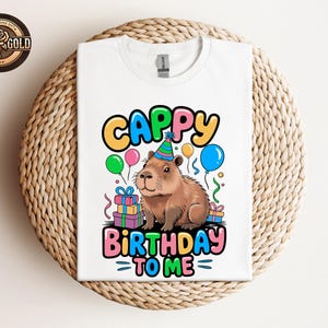 May include: White t-shirt with a colorful graphic featuring a capybara wearing a party hat, balloons, and the text "Cappy Birthday To Me". The design includes gift boxes and a circular logo in the upper left corner.