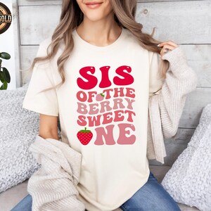 May include: Cream-colored t-shirt with the text "SIS OF THE BERRY SWEET ONE" in red and pink lettering, featuring strawberry graphics. The shirt has a round neckline and short sleeves. A "Rose Gold" logo is visible in the upper left corner.