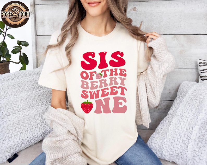 May include: Cream-colored t-shirt with the text "SIS OF THE BERRY SWEET ONE" in red and pink lettering, featuring strawberry graphics. The shirt has a round neckline and short sleeves. A "Rose Gold" logo is visible in the upper left corner.
