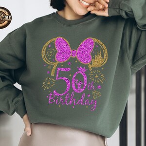 May include: A dark green sweatshirt with a glittery purple bow and "50th Birthday" text. The design includes gold accents of fireworks, a castle, and Mickey Mouse ears. The sweatshirt is a casual style.