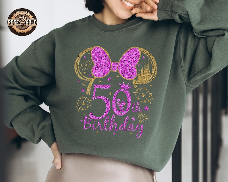 May include: A dark green sweatshirt with a glittery purple bow and "50th Birthday" text. The design includes gold accents of fireworks, a castle, and Mickey Mouse ears. The sweatshirt is a casual style.