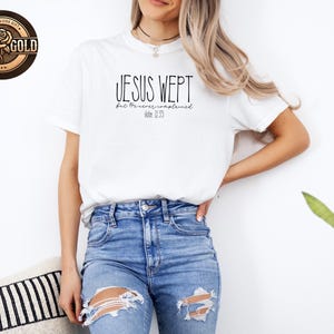 May include: White t-shirt with the text "JESUS WEPT" in black, with "but He never remained" and "John 11:35" below. The shirt is worn with blue jeans. A "Rose Gold" logo is visible in the upper left corner.