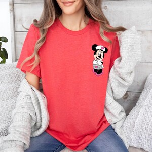 Puede incluir: Una camiseta de color coral con un gr&aacute;fico de Minnie Mouse. El personaje de dibujos animados lleva un gorro de enfermera y sostiene un regalo. La camiseta tiene mangas cortas y cuello redondo. El tema general es la ropa inspirada en Disney.