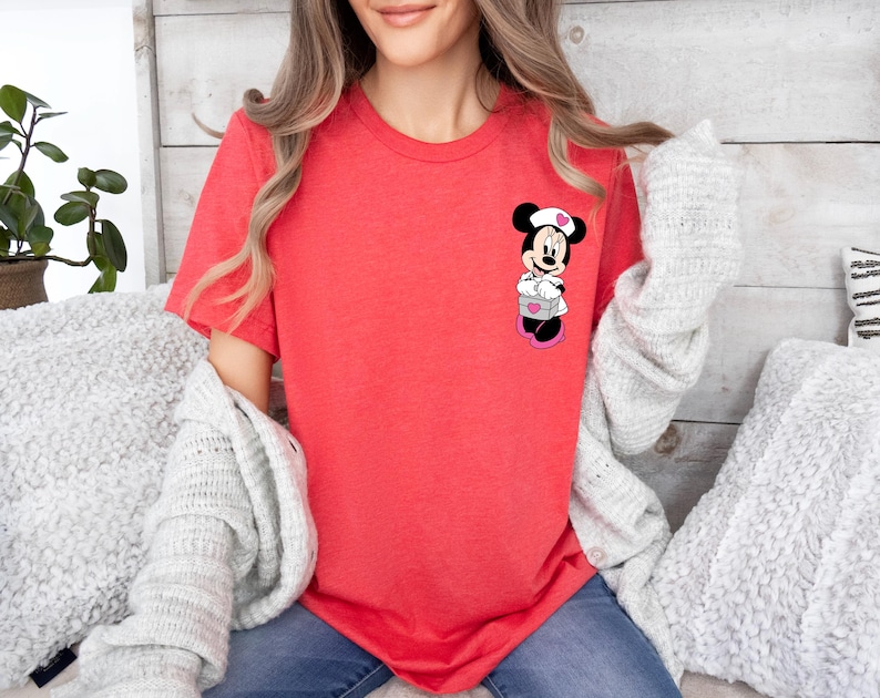 Puede incluir: Una camiseta de color coral con un gr&aacute;fico de Minnie Mouse. El personaje de dibujos animados lleva un gorro de enfermera y sostiene un regalo. La camiseta tiene mangas cortas y cuello redondo. El tema general es la ropa inspirada en Disney.
