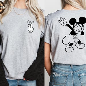 May include: A light grey t-shirt with a black and white Mickey Mouse design. The front displays the name "Taylor" above a peace sign hand gesture. The back of the shirt features Mickey Mouse doing a dab pose.