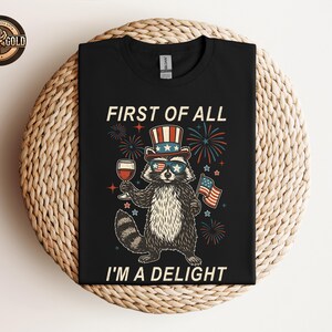 May include: Black t-shirt featuring a raccoon wearing sunglasses, a patriotic hat, and holding a glass of wine and an American flag. The text reads "FIRST OF ALL I'M A DELIGHT" with fireworks in the background.