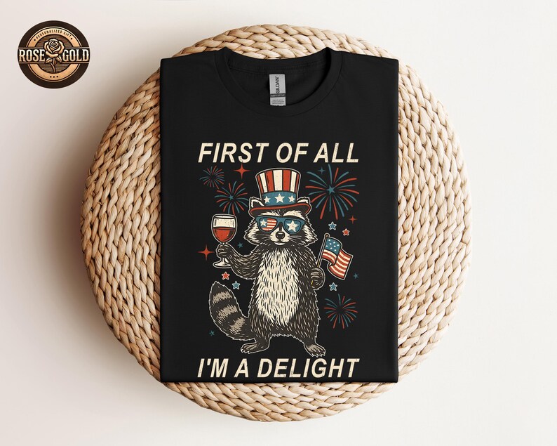 May include: Black t-shirt featuring a raccoon wearing sunglasses, a patriotic hat, and holding a glass of wine and an American flag. The text reads "FIRST OF ALL I'M A DELIGHT" with fireworks in the background.