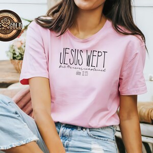May include: A light pink t-shirt with the text "JESUS WEPT but He never complained. John 11:35" in black. The shirt has short sleeves and a round neckline. A rose gold logo is visible in the upper left corner.