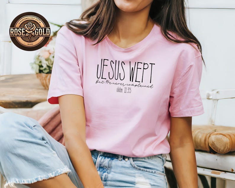 May include: A light pink t-shirt with the text "JESUS WEPT but He never complained. John 11:35" in black. The shirt has short sleeves and a round neckline. A rose gold logo is visible in the upper left corner.