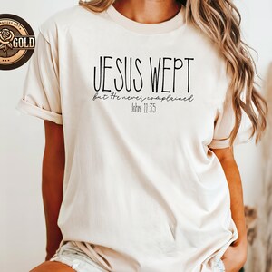 May include: A beige t-shirt with the text "JESUS WEPT but He never complained John 11:35" in black. The shirt has short sleeves and a relaxed fit. A rose gold sticker is visible in the upper left corner.