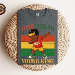 Puede incluir: Camiseta gris oscuro con una ilustraci&oacute;n de dibujos animados de un joven negro haciendo dab, con una corona, una camisa roja con "1865" y pantalones cortos amarillos. La camiseta tiene las palabras "Juneteenth" y "Young King" en naranja y verde.