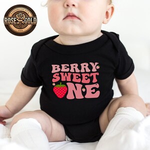 May include: Black baby bodysuit with the text "BERRY SWEET ONE" in pink and a red strawberry graphic. The bodysuit has short sleeves and is designed for a baby. The Rose Gold logo is in the upper left corner.