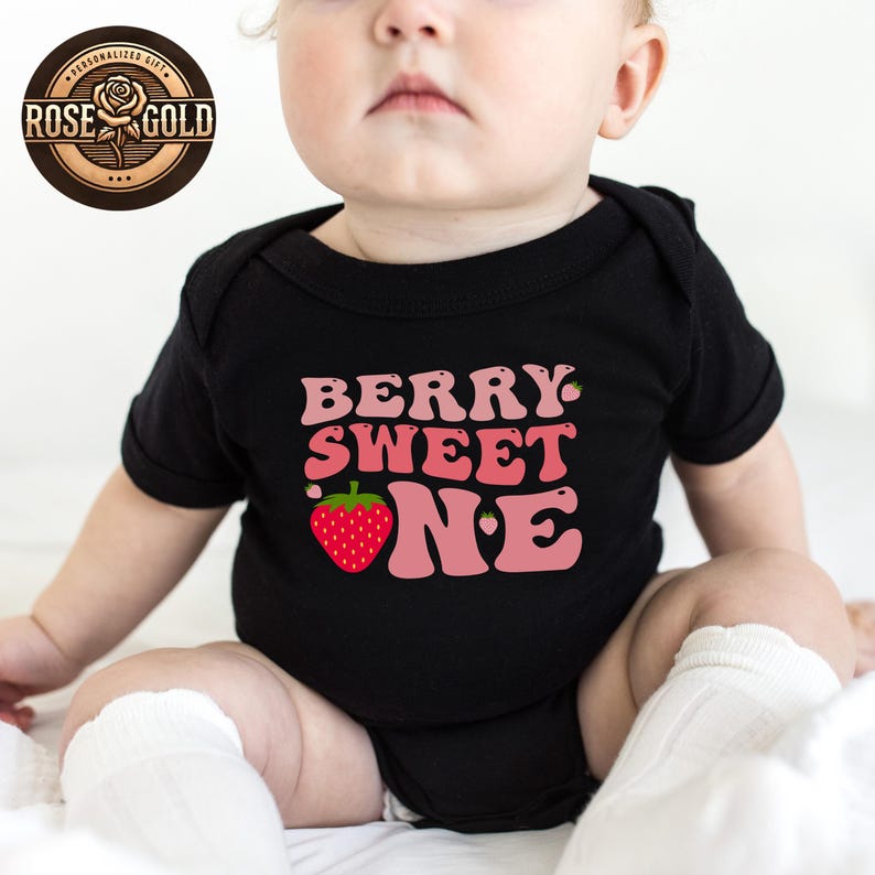 May include: Black baby bodysuit with the text "BERRY SWEET ONE" in pink and a red strawberry graphic. The bodysuit has short sleeves and is designed for a baby. The Rose Gold logo is in the upper left corner.