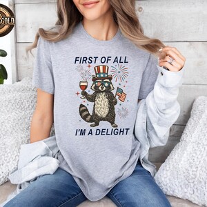 May include: A light gray t-shirt featuring a raccoon wearing sunglasses and a patriotic hat, holding a glass of wine and an American flag. The text reads "FIRST OF ALL I'M A DELIGHT" with fireworks in the background.