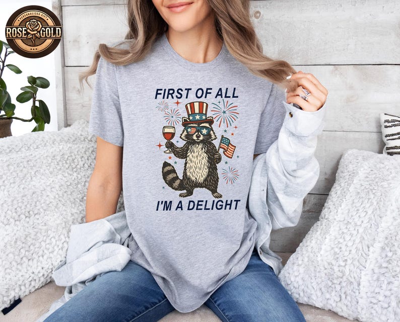 May include: A light gray t-shirt featuring a raccoon wearing sunglasses and a patriotic hat, holding a glass of wine and an American flag. The text reads "FIRST OF ALL I'M A DELIGHT" with fireworks in the background.