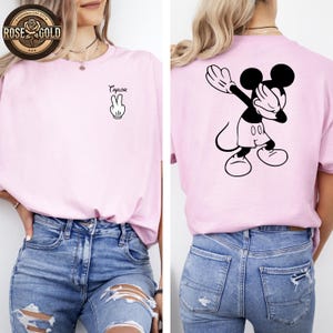 May include: A pale pink t-shirt with a black and white Mickey Mouse design on the back. The front of the shirt has the name "Taylor" above a peace sign. The shirt is worn with blue denim jeans.