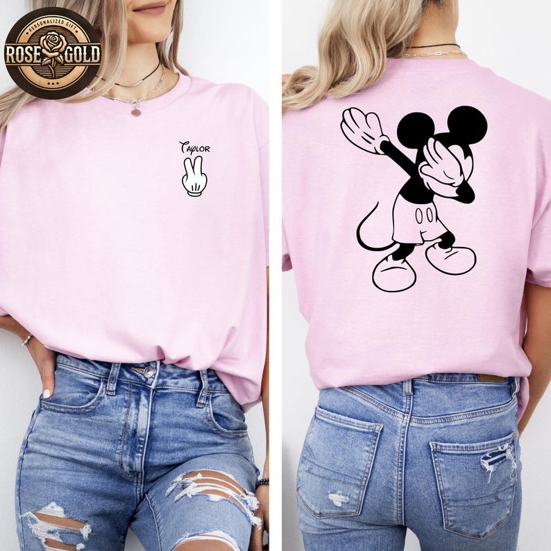 May include: A pale pink t-shirt with a black and white Mickey Mouse design on the back. The front of the shirt has the name "Taylor" above a peace sign. The shirt is worn with blue denim jeans.