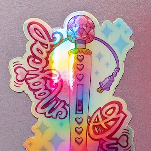 May include: Holographic sticker featuring a vibrator design with a faceted head, a body adorned with hearts, and the word "Love" in a stylized script. The sticker has a rainbow sheen and a plug.