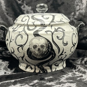 Peut inclure: Un sucrier en céramique blanche avec un motif de crâne et de serpent noir et blanc. Le bol a un couvercle et deux poignées.
