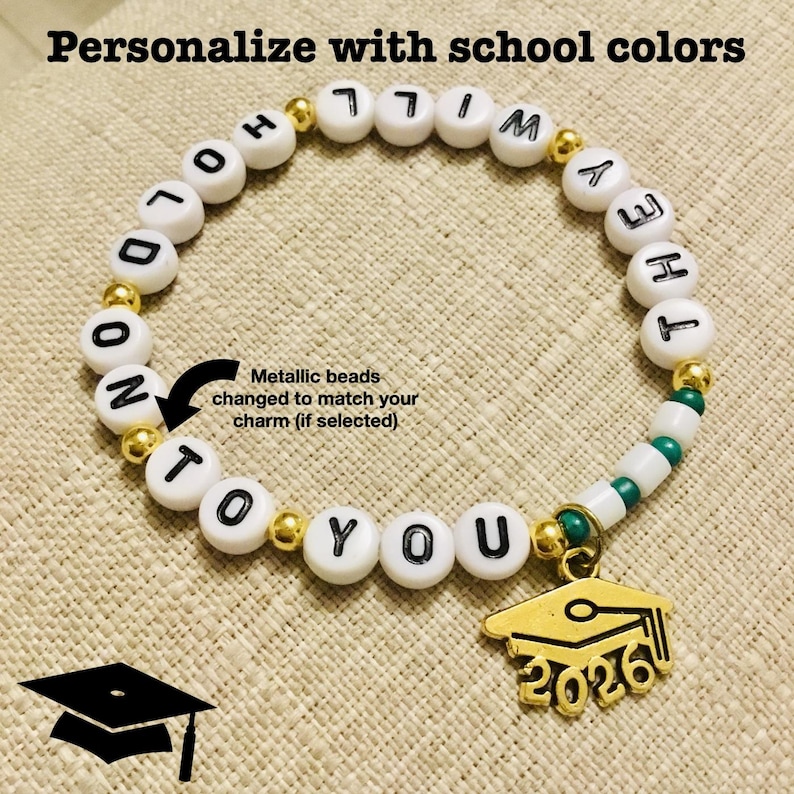 May include: A white beaded bracelet with black lettered beads spelling "HOLD ON TO YOU, Whitney Hey". The bracelet has gold metallic beads and a gold graduation cap charm with "2026" on it. The text "Personalise with school colours" is at the top.