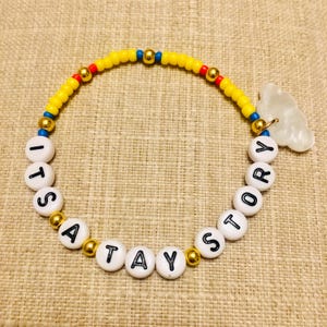 May include: A beaded bracelet featuring yellow, red, and blue seed beads interspersed with gold-tone metallic spheres. White circular beads with black lettering spell out ITS A TAY STORY, accompanied by a translucent white cloud-shaped charm.
