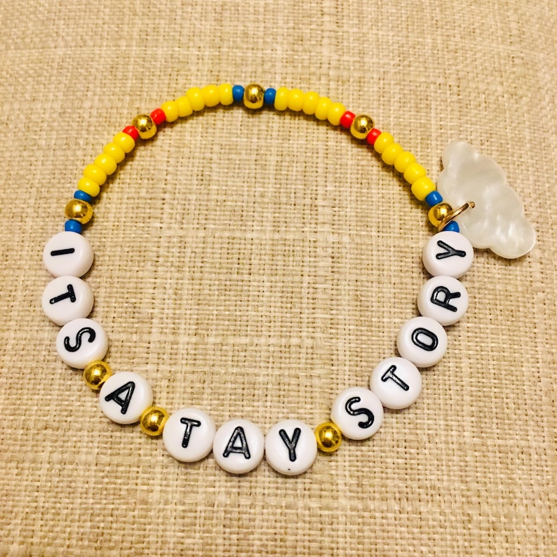 May include: A beaded bracelet featuring yellow, red, and blue seed beads interspersed with gold-tone metallic spheres. White circular beads with black lettering spell out ITS A TAY STORY, accompanied by a translucent white cloud-shaped charm.
