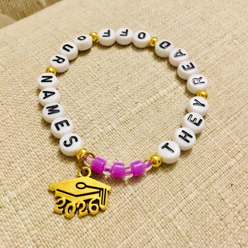 May include: A white beaded bracelet with black lettered beads spelling "OUR NAMES OFF THE READER THEY". A gold graduation cap charm with "2026" is attached. The bracelet also has gold and purple beads.