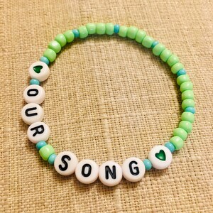 May include: A beaded bracelet with the words "OUR SONG" spelled out in black letters on white beads. The bracelet features green and blue beads, with heart-shaped accents. The bracelet is on a tan surface.