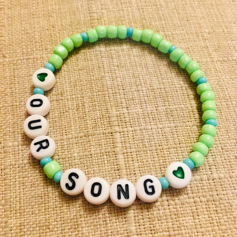 May include: A beaded bracelet with the words "OUR SONG" spelled out in black letters on white beads. The bracelet features green and blue beads, with heart-shaped accents. The bracelet is on a tan surface.