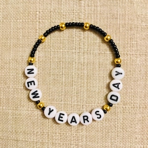 May include: A beaded bracelet with the words "NEW YEARS DAY" spelled out in white letter beads. The bracelet features black beads and gold-colored accents, perfect for celebrating the New Year.