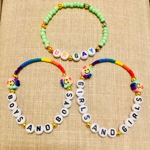 Op de afbeelding: Drie kralenarmbanden met kleurrijke plastic kralen en goudkleurige tussenstukjes. E&eacute;n armband heeft lichtgroene kralen met witte letterkralen die UR GAY spellen. Twee armbanden bevatten regenboogkleurige kralen, smiley-bedels en witte letterkralen die BOYS AND BOYS en GIRLS AND GIRLS spellen.