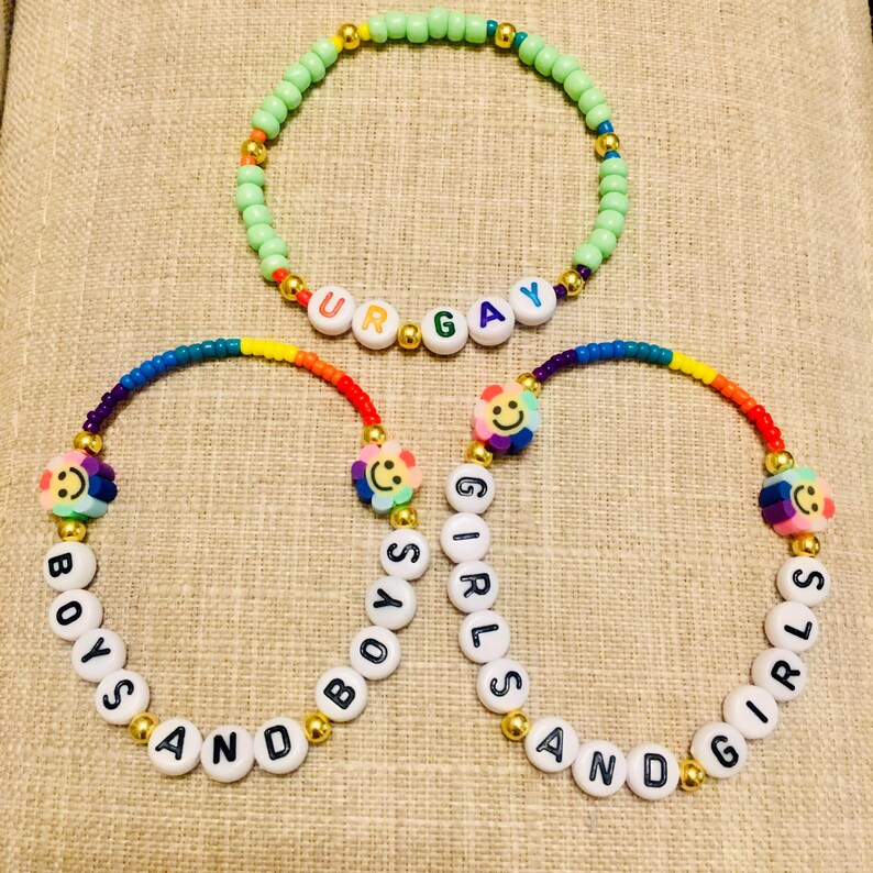 Op de afbeelding: Drie kralenarmbanden met kleurrijke plastic kralen en goudkleurige tussenstukjes. E&eacute;n armband heeft lichtgroene kralen met witte letterkralen die UR GAY spellen. Twee armbanden bevatten regenboogkleurige kralen, smiley-bedels en witte letterkralen die BOYS AND BOYS en GIRLS AND GIRLS spellen.