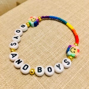Op de afbeelding: Een kralenarmband met witte ronde kralen met zwarte letters die BOYS AND BOYS spellen, afgewisseld met goudkleurige tussenkralen. Het ontwerp bevat twee veelkleurige bloemvormige kralen met smileygezichtjes en een regenboogkleurig segment.