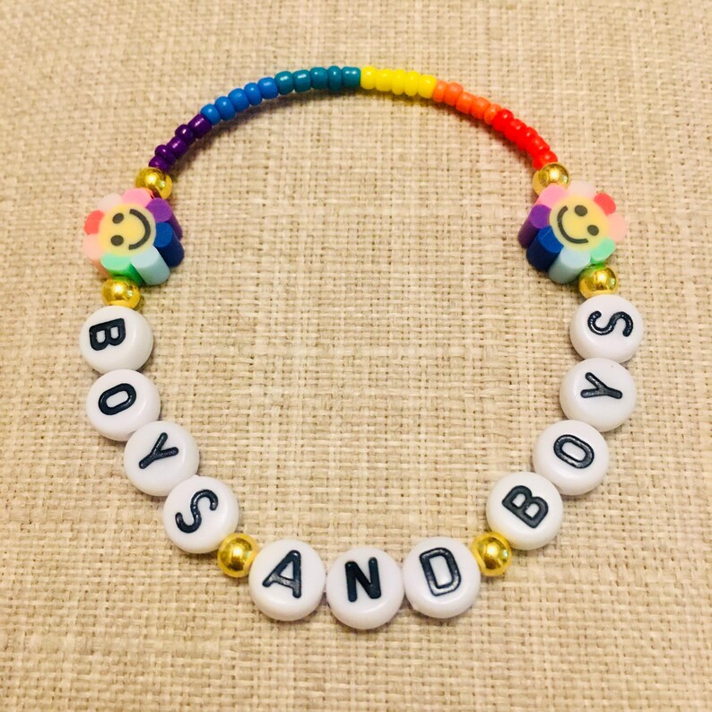 Op de afbeelding: Een kralenarmband met een regenboogkleurige boog van kleine kraaltjes, twee veelkleurige bloemvormige kralen met smileygezichtjes en witte ronde kralen met zwarte letters die de tekst BOYS AND BOYS vormen, geaccentueerd door goudkleurige kralen.