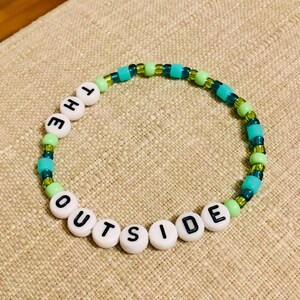 May include: A beaded bracelet with the words "THE OUTSIDE" spelled out in black letters on white beads. The bracelet also features turquoise, green, and clear beads, creating a colorful accessory.