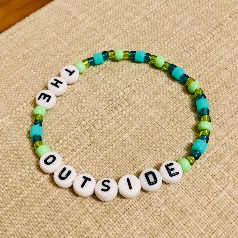 May include: A beaded bracelet with the words "THE OUTSIDE" spelled out in black letters on white beads. The bracelet also features turquoise, green, and clear beads, creating a colorful accessory.