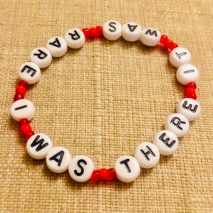 Puede incluir: Una pulsera circular de cuentas que alterna cuentas esf&eacute;ricas blancas con letras negras impresas y peque&ntilde;as cuentas espaciadoras rojas transl&uacute;cidas. Las cuentas blancas forman la frase I WAS THERE IT WAS RARE en una secuencia continua.