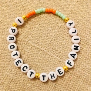May include: A white beaded bracelet with the words "I PROTECT THE FAMILY" in black letters. The bracelet has orange and green beads, with gold-colored accents.