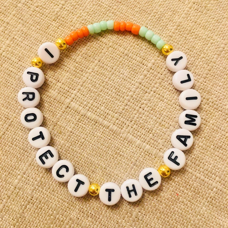 May include: A white beaded bracelet with the words "I PROTECT THE FAMILY" in black letters. The bracelet has orange and green beads, with gold-colored accents.