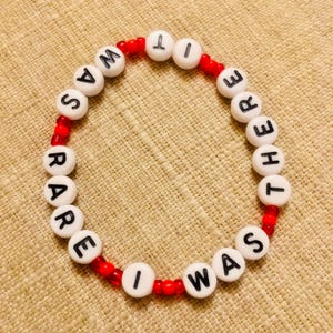 Puede incluir: Una pulsera circular de cuentas con cuentas redondas blancas que tienen letras negras impresas formando la frase IT WAS RARE I WAS THERE, intercaladas con peque&ntilde;as cuentas espaciadoras rojas transl&uacute;cidas en un patr&oacute;n.