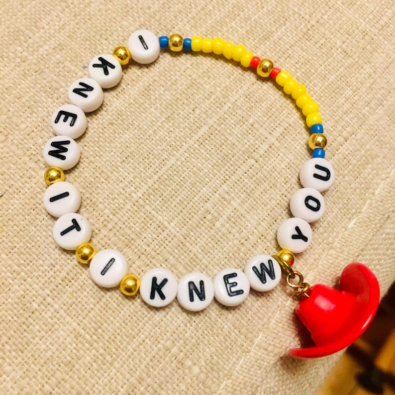 May include: A beaded bracelet featuring white round beads with black letters spelling I KNEW IT I KNEW YOU, interspersed with small gold metallic beads, a section of yellow beads, and a small red cowboy hat charm attached to the end of the band.