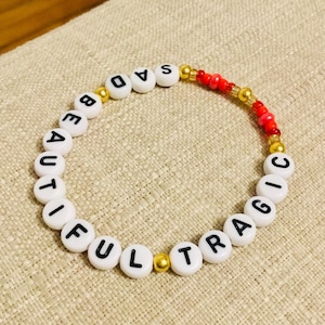 Puede incluir: Una pulsera circular de cuentas con abalorios redondos blancos y letras negras que forman la frase SAD BEAUTIFUL TRAGIC. El dise&ntilde;o incluye peque&ntilde;as cuentas espaciadoras de color dorado met&aacute;lico y una secci&oacute;n de cuentas rojas brillantes.
