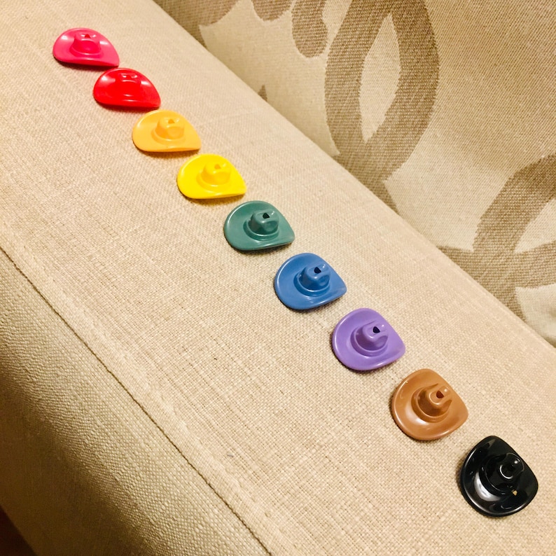 May include: A collection of ten miniature cowboy hats arranged in a diagonal line. The hats are colored in pink, red, orange, yellow, green, blue, purple, brown, and black, each featuring a small crown and a curved brim with a central hole.