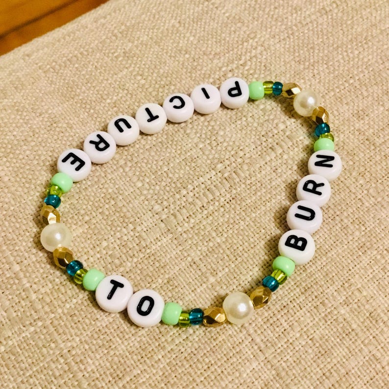 May include: A beaded bracelet with white letter beads spelling "PICTURE TO BURN". The bracelet features green, gold, and blue beads, along with faux pearls. The bracelet is on a beige fabric surface.
