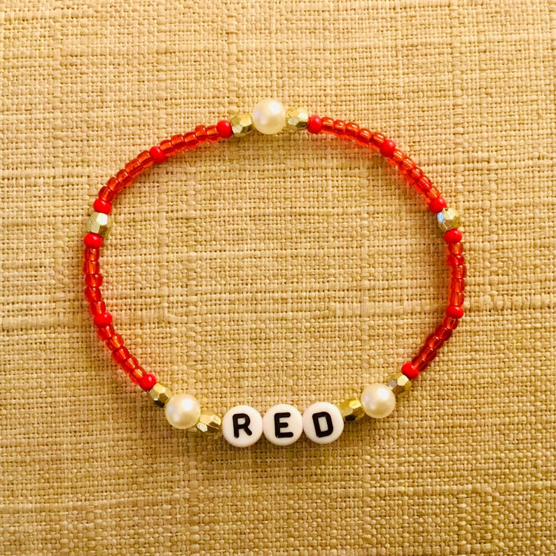 Puede incluir: Una pulsera de cuentas circular con peque&ntilde;as cuentas de vidrio rojo, espaciadores de metal dorado y dos cuentas blancas de imitaci&oacute;n perla. La pulsera incluye tres cuentas c&uacute;bicas blancas con letras negras que forman la palabra RED.