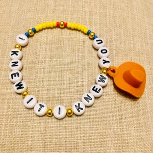 May include: A beaded bracelet featuring white round beads with black letters spelling out I KNEW IT I KNEW YOU, interspersed with small metallic gold, blue, red, and yellow beads, finished with an orange cowboy hat charm attached to the side.