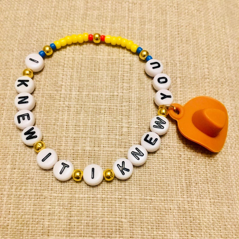 May include: A beaded bracelet featuring white round beads with black letters spelling out I KNEW IT I KNEW YOU, interspersed with small metallic gold, blue, red, and yellow beads, finished with an orange cowboy hat charm attached to the side.
