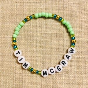 May include: A beaded bracelet with the name "TIM MCGRAW" spelled out in white letter beads. The bracelet features light green, gold, and teal beads, creating a colorful accessory.