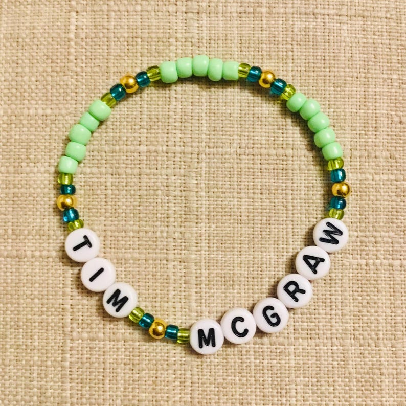 May include: A beaded bracelet with the name "TIM MCGRAW" spelled out in white letter beads. The bracelet features light green, gold, and teal beads, creating a colorful accessory.